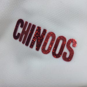 May include: Red glitter lettering on a white background that reads "CHINOOS".