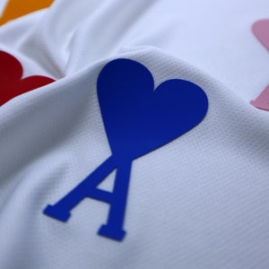 May include: A white fabric background with three heart-shaped patches. The patches are blue, pink, and red. Each patch has a stylized letter 'A' in the center.