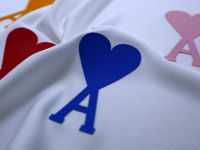 May include: A white fabric background with three heart-shaped patches. The patches are blue, pink, and red. Each patch has a stylized letter 'A' in the center.