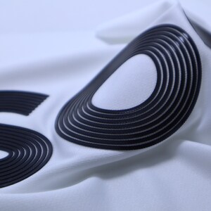 May include: Close-up of a white fabric with the word "SUPREME" printed in black raised lettering. The lettering is made of multiple layers of fabric, creating a 3D effect.
