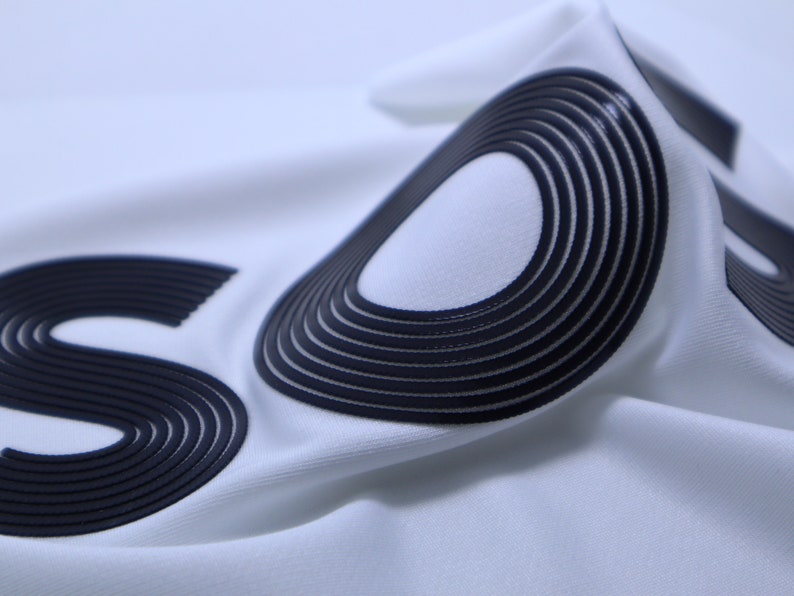 May include: Close-up of a white fabric with the word "SUPREME" printed in black raised lettering. The lettering is made of multiple layers of fabric, creating a 3D effect.