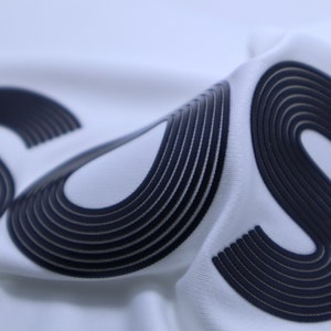 May include: Close-up of a white fabric with two black "S" letters. The letters are made of multiple layers of fabric, creating a 3D effect.