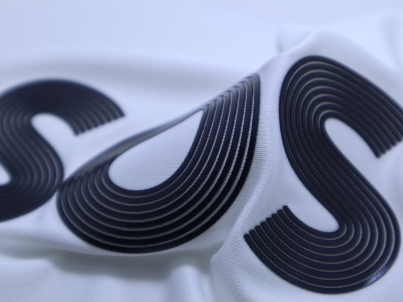 May include: Close-up of a white fabric with two black "S" letters. The letters are made of multiple layers of fabric, creating a 3D effect.