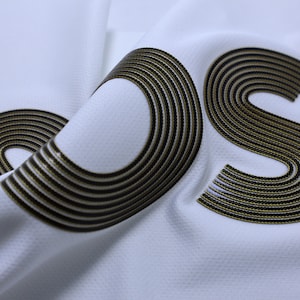 Pode incluir: Close-up de um tecido branco com um logotipo "OS" preto e dourado. O logotipo &eacute; feito de m&uacute;ltiplos c&iacute;rculos conc&ecirc;ntricos.
