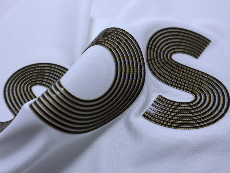 Pode incluir: Close-up de um tecido branco com um logotipo "OS" preto e dourado. O logotipo &eacute; feito de m&uacute;ltiplos c&iacute;rculos conc&ecirc;ntricos.