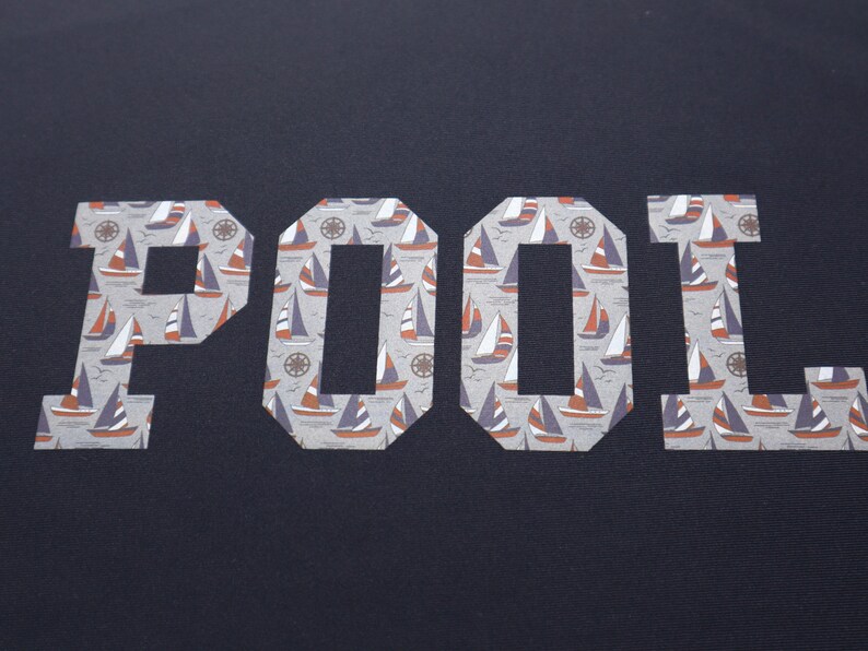 May include: A navy blue t-shirt with the word "POOL" printed in white letters. The letters are filled with a pattern of small sailboats.