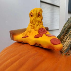 May include: A ceramic pizza slice with pepperoni and cheese topping, sitting on top of an orange pumpkin. The pizza slice has a cartoon face with googly eyes.