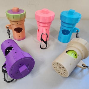 Op de afbeelding: Vijf verschillende gekleurde plastic containers met een gat aan de zijkant en een karabijnhaak. De containers zijn paars, roze, blauw, beige en groen en oranje ombre.