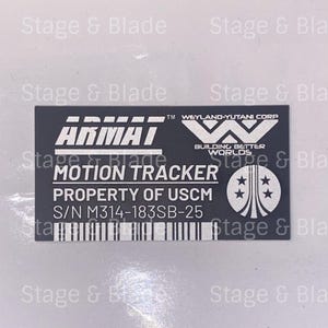 May include: A rectangular black and silver Armat Motion Tracker label. The label includes the text "ARMAI", "MOTION TRACKER", "PROPERTY OF USCM", and "S/N M314-183SB-25". The Weyland-Yutani Corp logo is also visible.