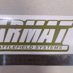 May include: A rectangular sticker with the text "ARMAI BATTLEFIELD SYSTEMS" in white and olive green. The sticker includes a "TM" symbol. A ruler is placed below the sticker.