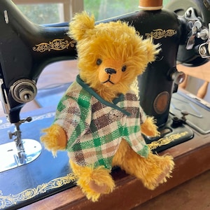 May include: A vintage-style teddy bear with golden fur and a plaid jacket in green, brown, and white. The bear is posed on a black antique sewing machine with gold detailing. The bear's jacket has a green collar and a small button closure.