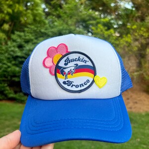 May include: A blue and white trucker hat with a yellow, red, and black patch that says "Buckin' Bronco" and a pink flower patch. There is a yellow heart patch on the side.