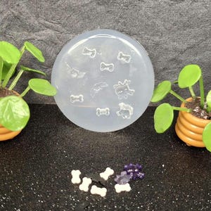 May include: A circular, translucent silicone mold featuring various small, recessed shapes including bones, bats, and a ghost. Two miniature potted plants with green leaves and orange ribbed pots flank the mold, with several small, finished resin pieces in white, black, and purple scattered in the foreground.