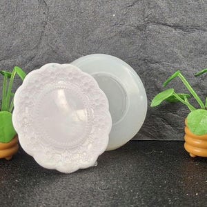 May include: A miniature scene featuring two small potted plants with bright green leaves and a white, decorative plate. The plants are in brown pots. A clear, round mold is also present, suggesting a crafting theme.