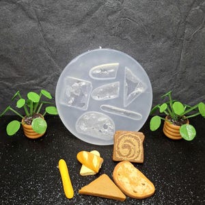 May include: A clear silicone mold with various bread shapes, including slices and a baguette, alongside miniature bread pieces. Two small potted plants flank the mold. The bread pieces are painted in shades of brown and yellow.