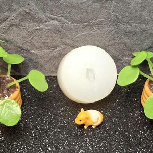 May include: A miniature scene featuring two small potted plants with bright green leaves and a white sphere. A tiny yellow and white guinea pig figurine is in the center. The plants are in orange pots.