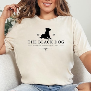 May include: A woman wearing a soft cream t-shirt with a black silhouette of a dog and the text "The Black Dog - Where Old Habits Die Screaming".