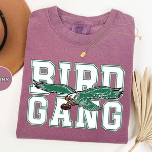 May include: A purple t-shirt with a white and green graphic of an eagle with the text "BIRD GANG" on it.