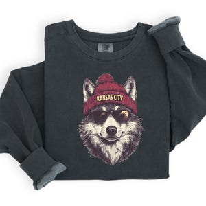 May include: A dark gray sweatshirt with a graphic of a wolf wearing a red beanie with the text "Kansas City" on it. The wolf is wearing sunglasses.