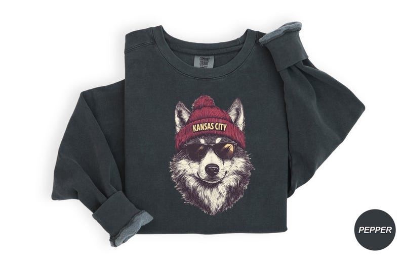 May include: A dark gray sweatshirt with a graphic of a wolf wearing a red beanie with the text "Kansas City" on it. The wolf is wearing sunglasses.