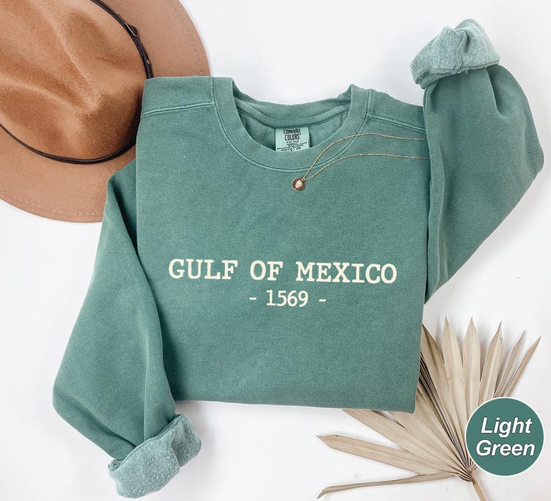 Op de afbeelding: Een lichtgroene sweatshirt met de witte tekst "GULF OF MEXICO - 1569 -". De sweatshirt heeft een ronde hals en lange mouwen met opgerolde manchetten. Een gouden ketting met een kleine hanger is over de voorkant gedrapeerd. Een bruine hoed is zichtbaar op de achtergrond.