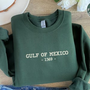 Op de afbeelding: Boszwarte sweatshirt met de witte tekst "GULF OF MEXICO - 1569 -". De mouwen zijn opgerold. Op de achtergrond is een witte hoed te zien. De sweatshirt is opgevouwen.