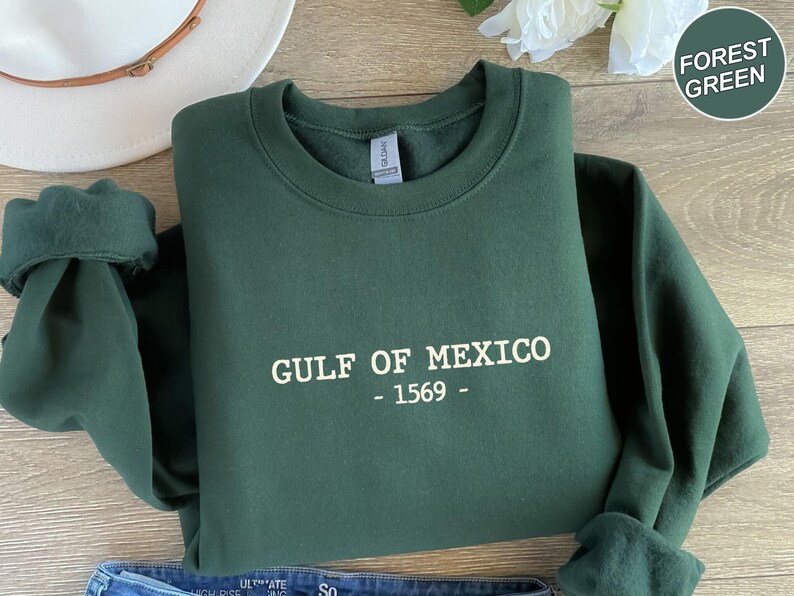 Op de afbeelding: Boszwarte sweatshirt met de witte tekst "GULF OF MEXICO - 1569 -". De mouwen zijn opgerold. Op de achtergrond is een witte hoed te zien. De sweatshirt is opgevouwen.