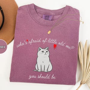 Pode incluir: Uma camiseta roxa com um gr&aacute;fico branco de um gato e o texto "who's afraid of little old me? you should be".