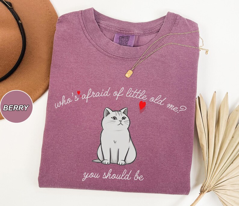 Pode incluir: Uma camiseta roxa com um gr&aacute;fico branco de um gato e o texto "who's afraid of little old me? you should be".