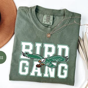 May include: A green t-shirt with a white graphic of an eagle with the words "Bird Gang" in white letters.