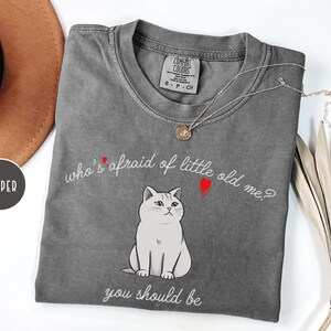 Pode incluir: Camiseta cinza com uma ilustra&ccedil;&atilde;o de um gato branco e o texto "who's afraid of little old me? you should be".