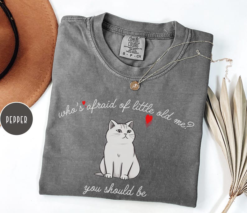 Pode incluir: Camiseta cinza com uma ilustra&ccedil;&atilde;o de um gato branco e o texto "who's afraid of little old me? you should be".