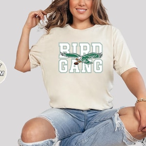 May include: A soft cream colored t-shirt with a green and white graphic of a bird with the text "Bird Gang" printed on the front.