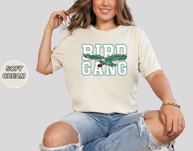 May include: A soft cream colored t-shirt with a green and white graphic of a bird with the text "Bird Gang" printed on the front.