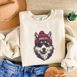 May include: A cream-colored sweatshirt with a graphic of a wolf wearing a red beanie with the text "Kansas City" on it. The wolf is wearing sunglasses and has a black nose and mouth.