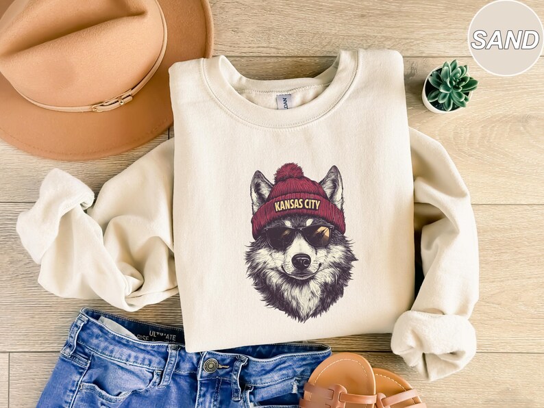 May include: A cream-colored sweatshirt with a graphic of a wolf wearing a red beanie with the text "Kansas City" on it. The wolf is wearing sunglasses and has a black nose and mouth.