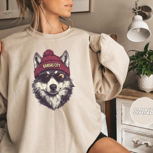 May include: A beige sweatshirt with a graphic of a wolf wearing a red beanie with the text "Kansas City" on it.