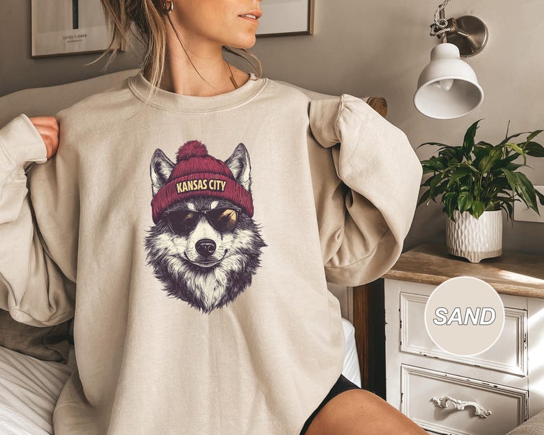 May include: A beige sweatshirt with a graphic of a wolf wearing a red beanie with the text "Kansas City" on it.