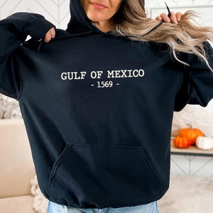 Op de afbeelding: Zwarte hoodie met de witte tekst "GULF OF MEXICO - 1569 -". De hoodie heeft een voorzak en een capuchon met trekkoord. Het woord "Black" staat in een cirkel linksonder.