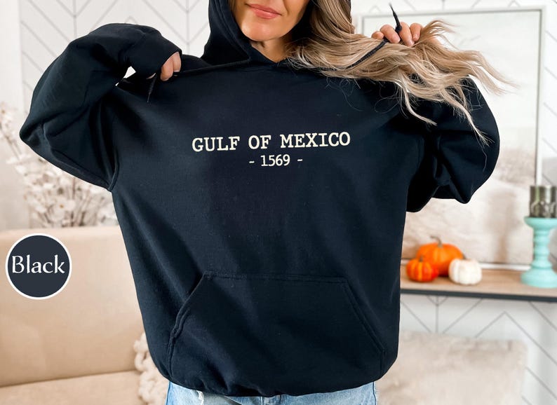 Op de afbeelding: Zwarte hoodie met de witte tekst "GULF OF MEXICO - 1569 -". De hoodie heeft een voorzak en een capuchon met trekkoord. Het woord "Black" staat in een cirkel linksonder.