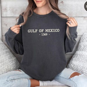 Op de afbeelding: Donkergrijs sweatshirt met de witte tekst "GULF OF MEXICO - 1569". Het sweatshirt is een crew neck stijl en wordt door een persoon gedragen. De persoon draagt lichtblauwe jeans met scheuren.