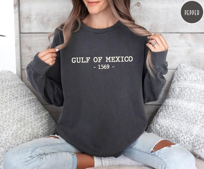 Op de afbeelding: Donkergrijs sweatshirt met de witte tekst "GULF OF MEXICO - 1569". Het sweatshirt is een crew neck stijl en wordt door een persoon gedragen. De persoon draagt lichtblauwe jeans met scheuren.
