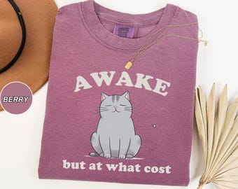 May include: A purple t-shirt with a gray cat illustration and the text "Awake but at what cost".
