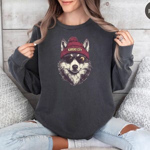 May include: A dark gray sweatshirt with a graphic of a wolf wearing a red beanie with the text "Kansas City" on it. The wolf is wearing sunglasses and has a playful expression.