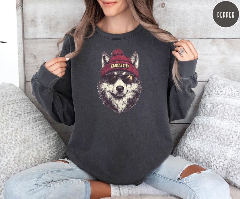 May include: A dark gray sweatshirt with a graphic of a wolf wearing a red beanie with the text "Kansas City" on it. The wolf is wearing sunglasses and has a playful expression.