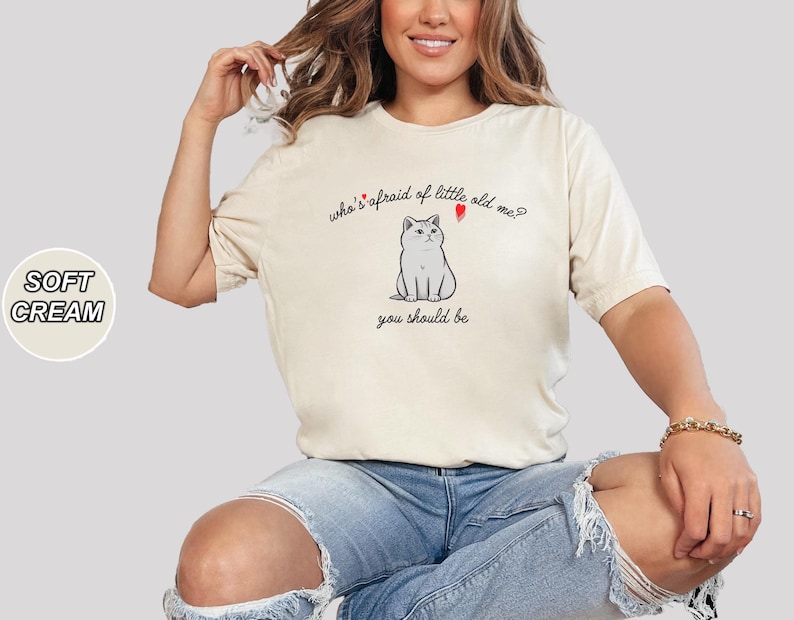 Pode incluir: Uma mulher vestindo uma camiseta creme com o texto "who's afraid of little old me?" e uma imagem de um gato de desenho animado. O texto "you should be" est&aacute; abaixo da imagem. Ela est&aacute; usando jeans azul.