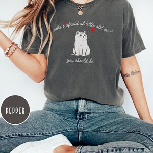 Pode incluir: Uma camiseta cinza escura com uma ilustra&ccedil;&atilde;o de um gato branco e o texto "who's afraid of little old me? you should be".