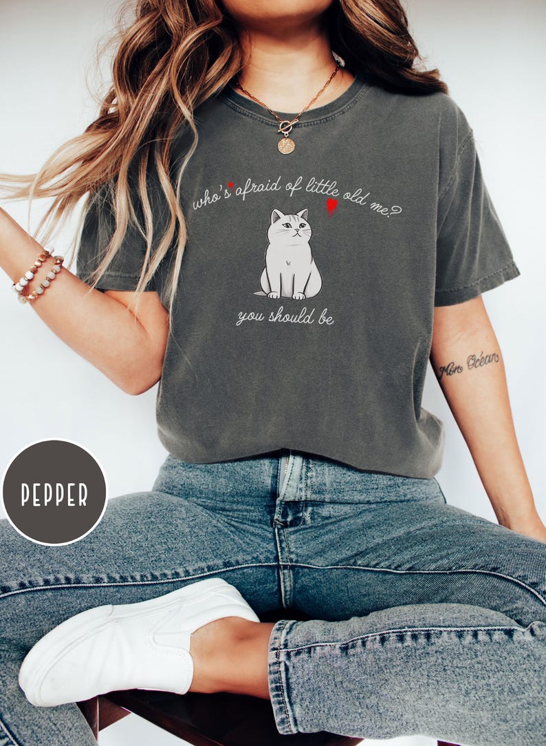 Pode incluir: Uma camiseta cinza escura com uma ilustra&ccedil;&atilde;o de um gato branco e o texto "who's afraid of little old me? you should be".
