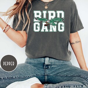 May include: A dark green t-shirt with the words "Bird Gang" in white lettering. A green and white eagle graphic is above the text. The shirt is being worn by a person.