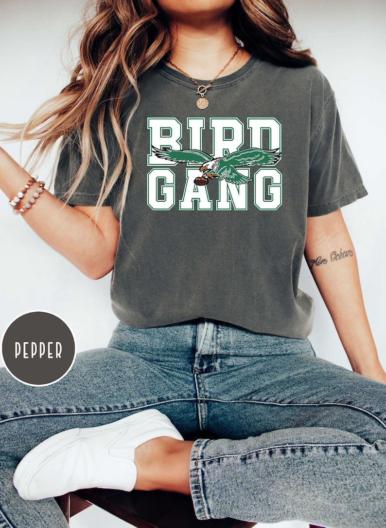 May include: A dark green t-shirt with the words "Bird Gang" in white lettering. A green and white eagle graphic is above the text. The shirt is being worn by a person.